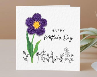 May include: A greeting card with a crocheted purple flower with a yellow center. The card has the text "HAPPY Mother's Day" in black script. Green leaves and stems support the flower, with delicate black line art of flowers at the bottom.