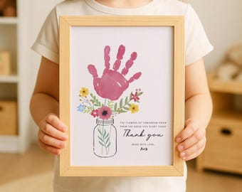 May include: A framed print featuring a handprint in pink, with a floral arrangement emerging from a mason jar. The print includes the text "Thank you" and "Made with love, Ava". The frame is made of light wood.
