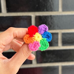May include: A vibrant, crocheted flower with petals in red, orange, yellow, pink, green, blue, and purple. The handmade flower is held in a hand, displaying its rainbow colour scheme. The flower is a craft item.