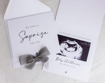 May include: A white pregnancy announcement card with the text "WE HAVE A Surprise FOR YOU" and a gray ribbon. The inside reveals an ultrasound image and the text "YOU'RE GOING TO BE GRANDPARENTS!" with the baby's name and due date.