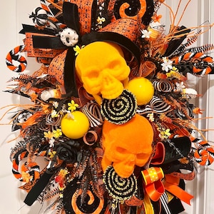 May include: A decorative Halloween wreath featuring two skulls, one yellow and one orange, surrounded by black and orange ribbons, skeleton-patterned fabric, black floral accents, yellow spheres, and black and orange spiral ornaments.