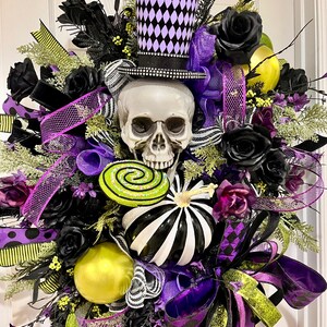 May include: A decorative wreath featuring a central human skull wearing a purple and black diamond-patterned top hat. The arrangement includes black roses, a black and white striped pumpkin, a lime green swirl lollipop, and purple ribbons.