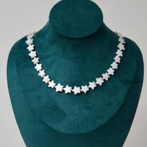 May include: A star-shaped necklace displayed on a green velvet bust. The necklace features white star pendants strung together with gold-colored links. The background is a solid, neutral color.