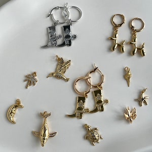 May include: A collection of gold and silver earrings featuring various charms, including a hummingbird, a palm tree, a crescent moon, a whale, a turtle, a cowboy boot, a balloon animal, and a starfish.