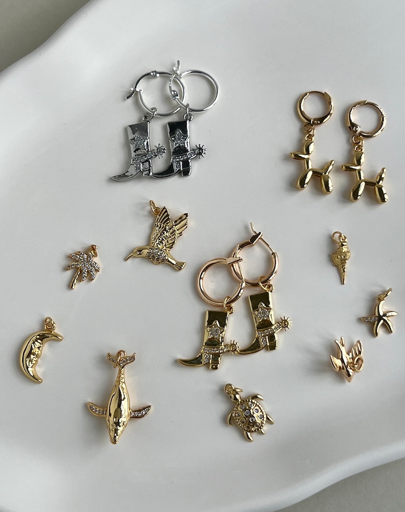 May include: A collection of gold and silver earrings featuring various charms, including a hummingbird, a palm tree, a crescent moon, a whale, a turtle, a cowboy boot, a balloon animal, and a starfish.