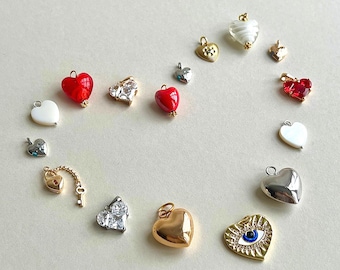 May include: A collection of various heart-shaped charms and pendants in gold and silver tones. The assortment includes red glass hearts, white stone hearts, clear crystal hearts, a gold heart with an evil eye design, and a small gold lock and key charm.