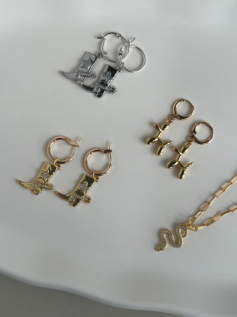 May include: A set of gold and silver jewelry featuring cowboy boot charms, balloon animal charms, and a snake charm. The jewelry includes earrings and a necklace.