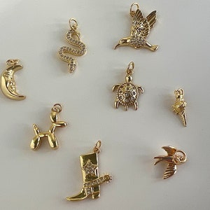 May include: A set of nine gold-tone charms featuring various designs, including a crescent moon, a snake, a hummingbird, a turtle, a balloon animal, a cowboy boot, and a bird. The charms are all detailed and have a sparkly finish.
