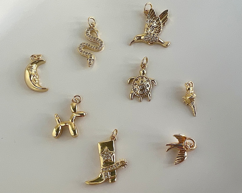 May include: A set of nine gold-tone charms featuring various designs, including a crescent moon, a snake, a hummingbird, a turtle, a balloon animal, a cowboy boot, and a bird. The charms are all detailed and have a sparkly finish.