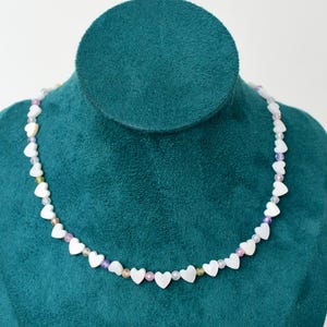 Puede incluir: Un delicado collar con cuentas blancas en forma de corazón intercaladas con pequeñas cuentas de colores. El collar se exhibe sobre un busto de terciopelo verde azulado, mostrando su diseño y artesanía. Una joya encantadora.