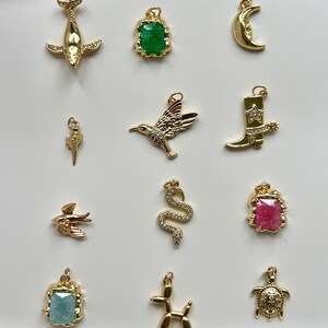 May include: A collection of 12 gold-tone charms featuring various designs, including a whale, a hummingbird, a cowboy boot, a snake, a turtle, a fish, a dog, a crescent moon, and three square charms with green, pink, and blue gemstones.