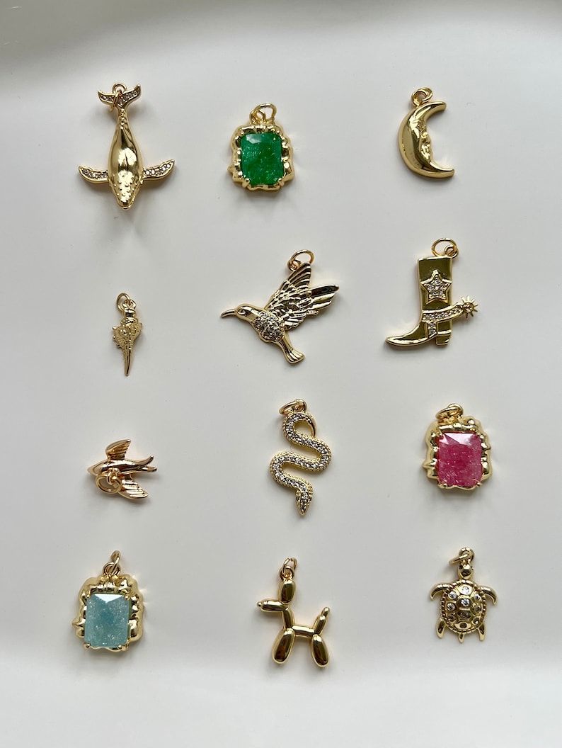 May include: A collection of 12 gold-tone charms featuring various designs, including a whale, a hummingbird, a cowboy boot, a snake, a turtle, a fish, a dog, a crescent moon, and three square charms with green, pink, and blue gemstones.