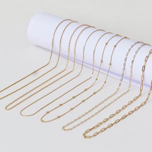 Könnte beinhalten: Eine Sammlung von goldenen Kettenhalsketten in verschiedenen Längen und Stilen, darunter eine Perlenkette, eine Gliederkette und eine Panzerkette. Die Ketten sind auf einer weißen Oberfläche angeordnet.