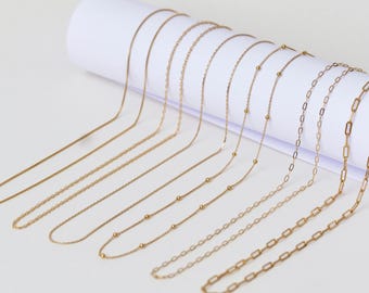 May include: A collection of gold chain necklaces of varying lengths and styles, including a beaded chain, a box chain, and a cable chain. The chains are arranged on a white surface.