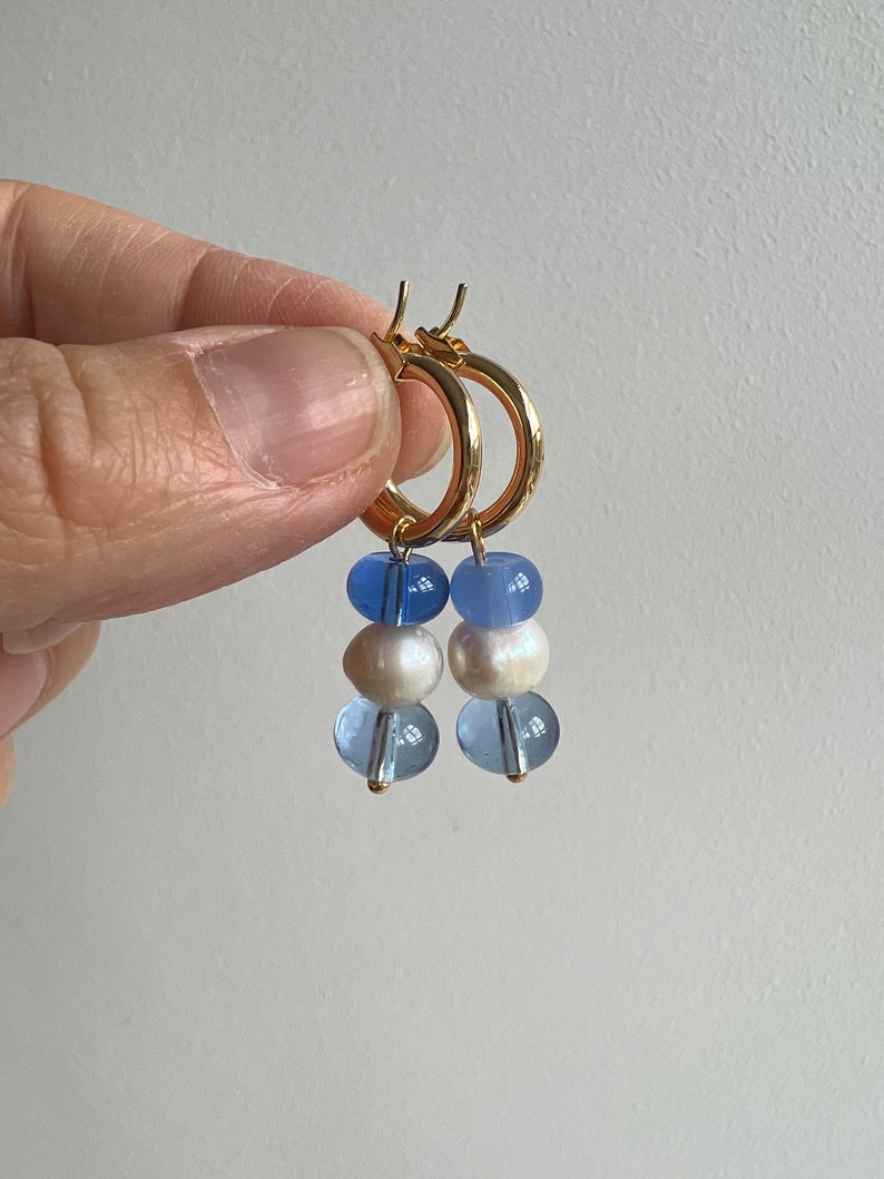 Pode incluir: Brincos de argola dourados com tr&ecirc;s contas azuis e brancas penduradas em cada argola.
