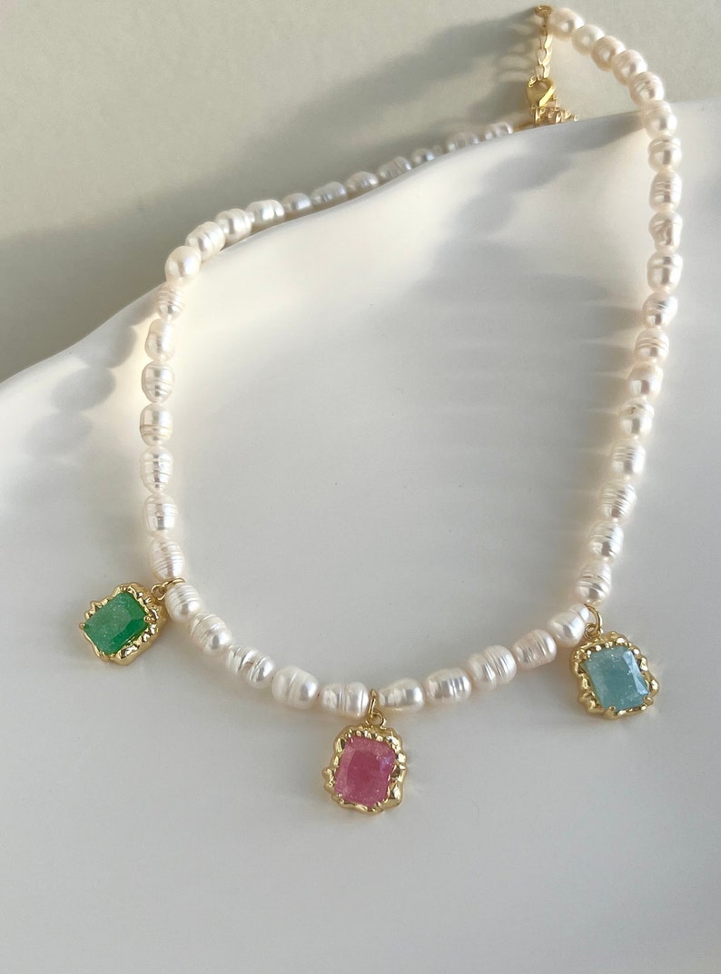May include: A delicate pearl necklace with three dangling charms. The charms are square-shaped and feature green, pink, and blue gemstones.