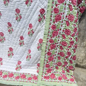 May include: A white quilted blanket featuring a repeating pattern of small pink rose clusters. The border displays a dense floral print of pink roses with green leaves and a thin light green striped edge, creating a distinct visual contrast.