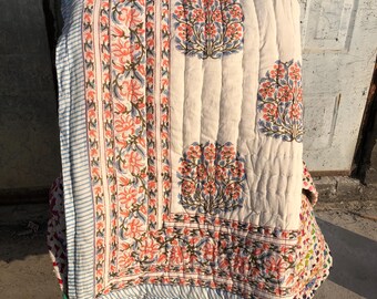 May include: A cream-colored quilt with a floral pattern in shades of coral, blue, and green. The quilt features a striped border and a repeating tree-like design. The quilt is folded over a chair.