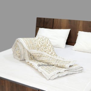 Könnte beinhalten: Eine aufgerollte, cremefarbene Steppdecke mit floralem Muster und grauem Rand liegt auf einem weißen Bett. Die Steppdecke ist teilweise entfaltet und zeigt ihr Design. Der Text "QuiltHubbazar" ist auf der Steppdecke zu sehen. Zwei weiße Kissen und ein Holz-Kopfteil sind im Hintergrund.