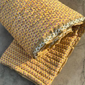 Puede incluir: Una manta acolchada con un patrón repetitivo de pequeños motivos florales rojos y verdes sobre un fondo amarillo brillante. El tejido tiene un borde liso de color tostado y líneas de costura visibles que crean una textura de cuadrícula.