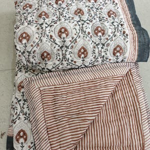 Pode incluir: Um edredom dobrado com fundo branco e um padrão repetido de motivos florais castanhos e cinzentos. O verso do edredom apresenta riscas castanhas e brancas. As bordas são finalizadas com uma borda cinza escuro.