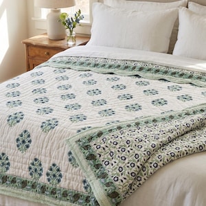 Puede incluir: Un edredón blanco con un estampado floral en tonos azules y verdes, colocado sobre una cama. El edredón presenta un diseño floral repetido y un borde decorativo. La cama está hecha con sábanas y almohadas blancas, creando una escena de dormitorio serena y acogedora.