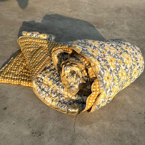Op de afbeelding: Een opgerolde quilt en een gedeeltelijk uitgevouwen quilt liggen op een betonnen oppervlak. De quilt heeft een geel en wit bloemenpatroon en een geel en wit geruit patroon. De randen zijn afgewerkt met een effen gele rand.