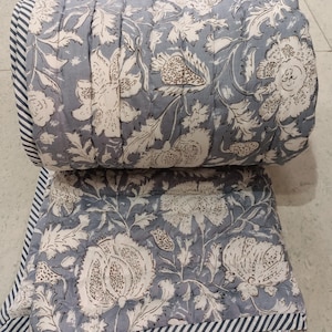 Può includere: Un piumone con motivo floreale grigio e bianco con un bordo a righe. Il piumone è piegato a metà e si trova su un pavimento di piastrelle bianche.