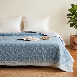 Peut inclure: Un couvre-lit matelassé bleu et blanc avec un motif floral répétitif et une bordure décorative. Le couvre-lit est posé sur un lit avec deux oreillers blancs. Un livre ouvert et une petite assiette de nourriture sont placés au centre du lit.