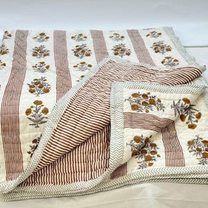 Op de afbeelding: Een wit en bruin gestreept katoenen dekbed met een bloemenprint. Het dekbed is dubbelgevouwen en heeft een decoratieve rand.