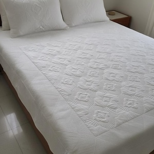 May include: A solid white bedspread featuring a central panel with a repeating quilted floral pattern and a wide border with linear stitching. Two matching white quilted pillow shams with similar floral motifs are positioned at the head of the bed.