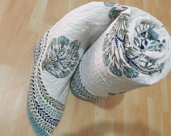 May include: A white quilted blanket with a floral pattern in shades of green and blue. The blanket is rolled up and has a decorative border.