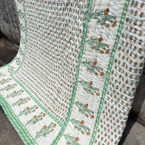 May include: A white quilted blanket featuring a repeating pattern of small orange flowers and green stems. The design includes a wide border with larger orange floral motifs and green geometric accents, all finished with a visible stitched texture.