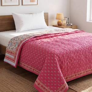 Op de afbeelding: Een roze gewatteerde sprei met een bloemenpatroon en decoratieve rand. De sprei ligt op een bed met een wit kussen en een houten hoofdbord. De sprei is ongeveer 203 cm lang en 152 cm breed.
