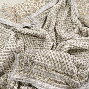 May include: A folded, off-white quilt featuring a repeating floral pattern in muted olive green. The quilt has a border with a geometric design and a light grey zigzag edge. The fabric appears soft and comfortable.
