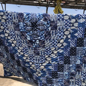 Könnte beinhalten: Eine große, handgefertigte Steppdecke mit Patchwork-Design. Die Steppdecke zeigt verschiedene Blau- und Weißtöne mit unterschiedlichen Mustern, darunter Punkte, Streifen und geometrische Formen. Die Steppdecke hängt im Freien.