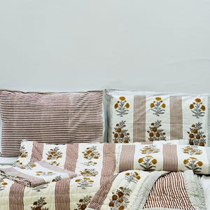 May include: A set of striped bedding with a floral print. The bedding includes a quilt, two pillowcases, and a pillow. The quilt and pillowcases are white with red stripes and a floral print in shades of brown and yellow. The pillow is white with red stripes.