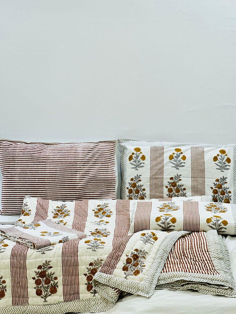 May include: A set of striped bedding with a floral print. The bedding includes a quilt, two pillowcases, and a pillow. The quilt and pillowcases are white with red stripes and a floral print in shades of brown and yellow. The pillow is white with red stripes.