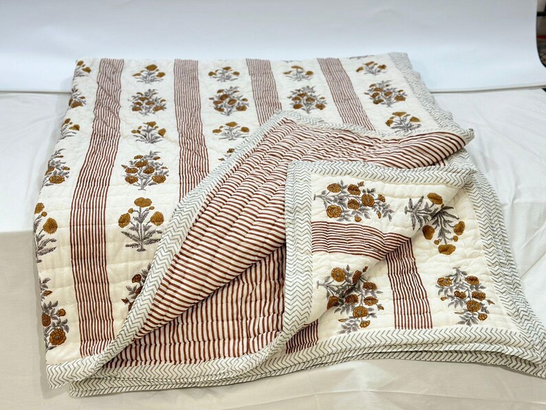 May include: A white and brown striped cotton quilt with a floral print. The quilt is folded in half and has a decorative border.