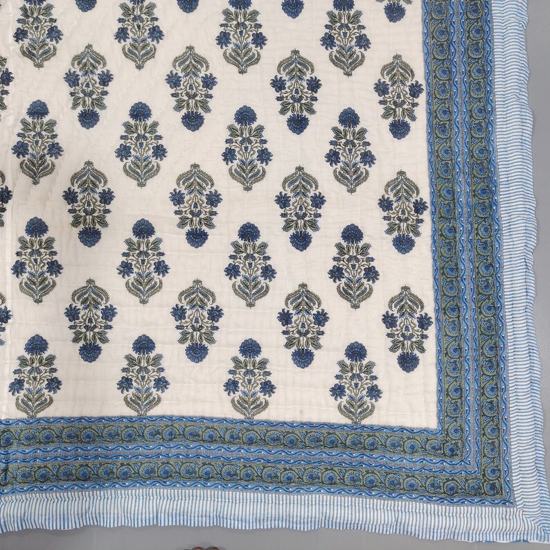 Op de afbeelding: Een wit katoenen dekbed met een blauw en groen bloemenpatroon. Het dekbed heeft een blauwe en groene rand met een witte streep.