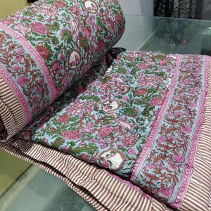 Op de afbeelding: Een blauwe en roze quilt met een bloemenpatroon en een witte rand. De quilt is dubbelgevouwen en heeft een gestreept patroon aan de achterkant.
