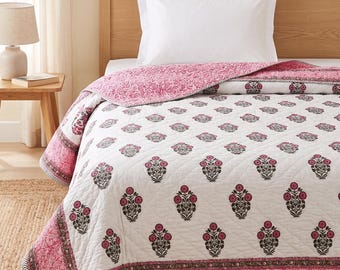May include: A white quilted bedspread featuring a repeating pattern of dark gray and pink floral motifs. The quilt has a wide pink border with a detailed paisley print and a thin black and white chevron edge, draped over a bed with a white pillow.