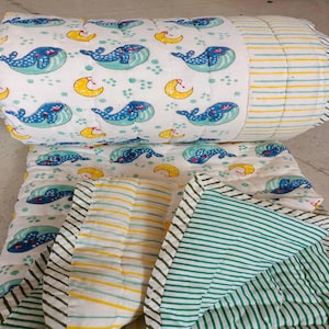Può includere: Un set di trapuntino e cuscino a rullo per bambini bianchi con un motivo a balena e mezzaluna in blu, verde acqua e giallo. La trapunta ha un bordo a righe verdi e bianche.