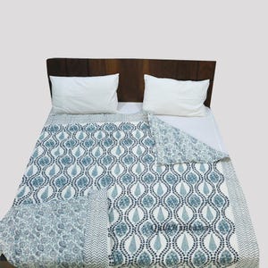 May include: A white and blue patterned cotton quilt with a floral design on the reverse side. The quilt is folded on a bed with white pillowcases and a wooden headboard.