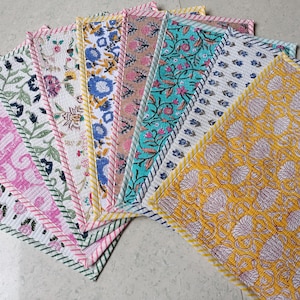 Op de afbeelding: Een collectie rechthoekige gewatteerde placemats gerangschikt in een waaier. Elke placemat heeft een uniek bloemenpatroon met blokdruk in diverse kleuren zoals geel, turquoise, roze, blauw en groen, afgewerkt met stoffen randen met strepen.