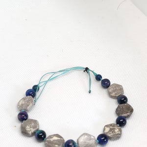 May include: A bracelet made with grey, hexagonal beads and smaller, dark blue and purple beads. The bracelet is strung on a light blue cord, with a simple knot closure.