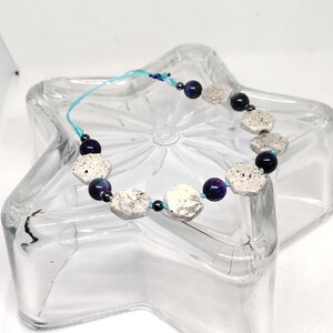 May include: A handcrafted bracelet with white and grey stone beads, dark blue and purple beads, and a light blue cord. The bracelet is presented on a clear glass star-shaped dish. The beads are likely around 1 cm in diameter.