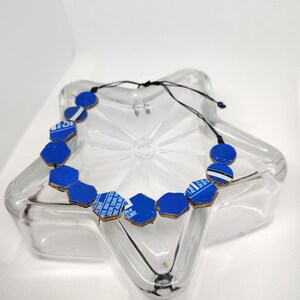 May include: A blue and brown necklace with geometric shapes. The necklace features blue hexagonal and circular beads with a brown border, strung on a black cord. The necklace is displayed on a clear, star-shaped glass dish.