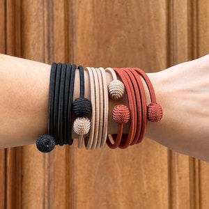 Puede incluir: Una variedad de pulseras en negro, beige y rojo óxido. Cada pulsera presenta un diseño enrollado y está adornada con una bola decorativa. Las pulseras se muestran en un brazo, mostrando sus colores y texturas.