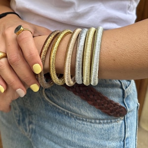 Puede incluir: Seis pulseras metálicas en tonos dorados, plateados y bronce. Las pulseras están apiladas en un brazo, con un cinturón trenzado marrón y vaqueros azules en el fondo. Las pulseras tienen un aspecto texturizado y tejido.